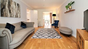 A living room with a beige sofa, a patterned rug, a blue armchair, and a TV stand. - Morrison House Holytown (Motherwell)