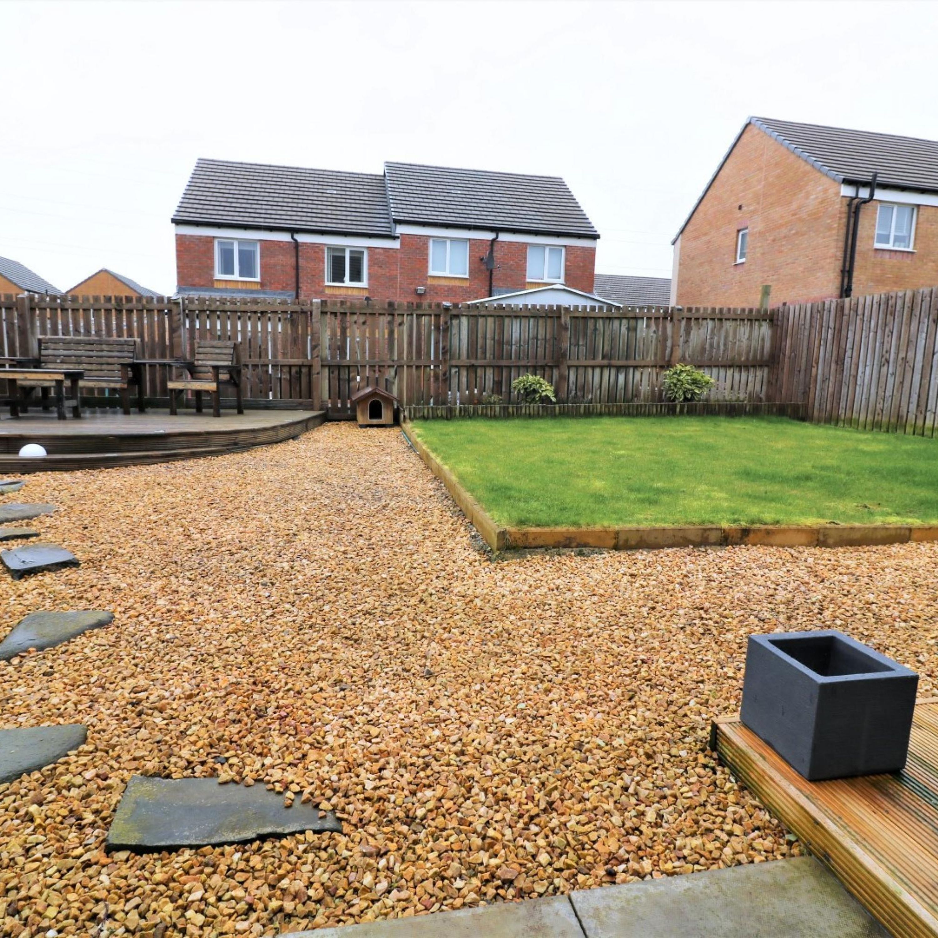 A backyard with a gravel path, a wooden deck, a grassy lawn, and a wooden fence.