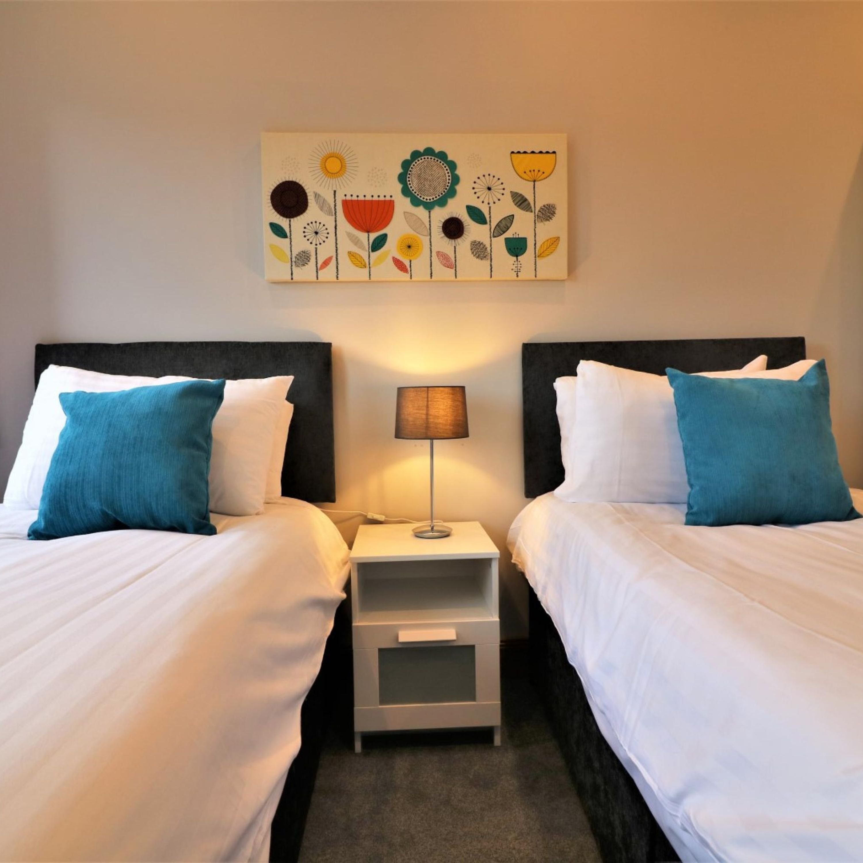 A hotel room with two beds, each with white bedding and teal pillows. There is a small bedside table with a lamp between the beds. Above the beds, there is a colorful artwork featuring flowers and leaves.