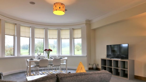 Een moderne woonkamer met een gebogen plafond, een groot raam met uitzicht op het omliggende landschap, een flatscreen televisie op een standaard en een eethoek met een tafel gedekt voor twee personen.