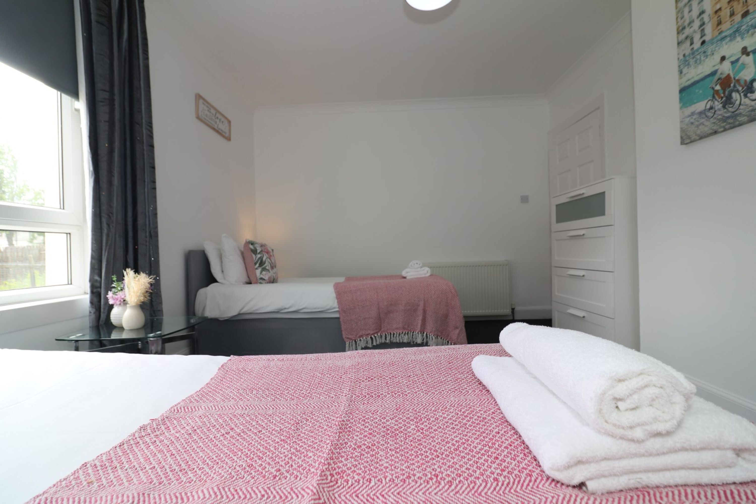 Una habitación de hotel con cama, una mesita de noche con lámpara, una ventana con cortinas y un cuadro en la pared.