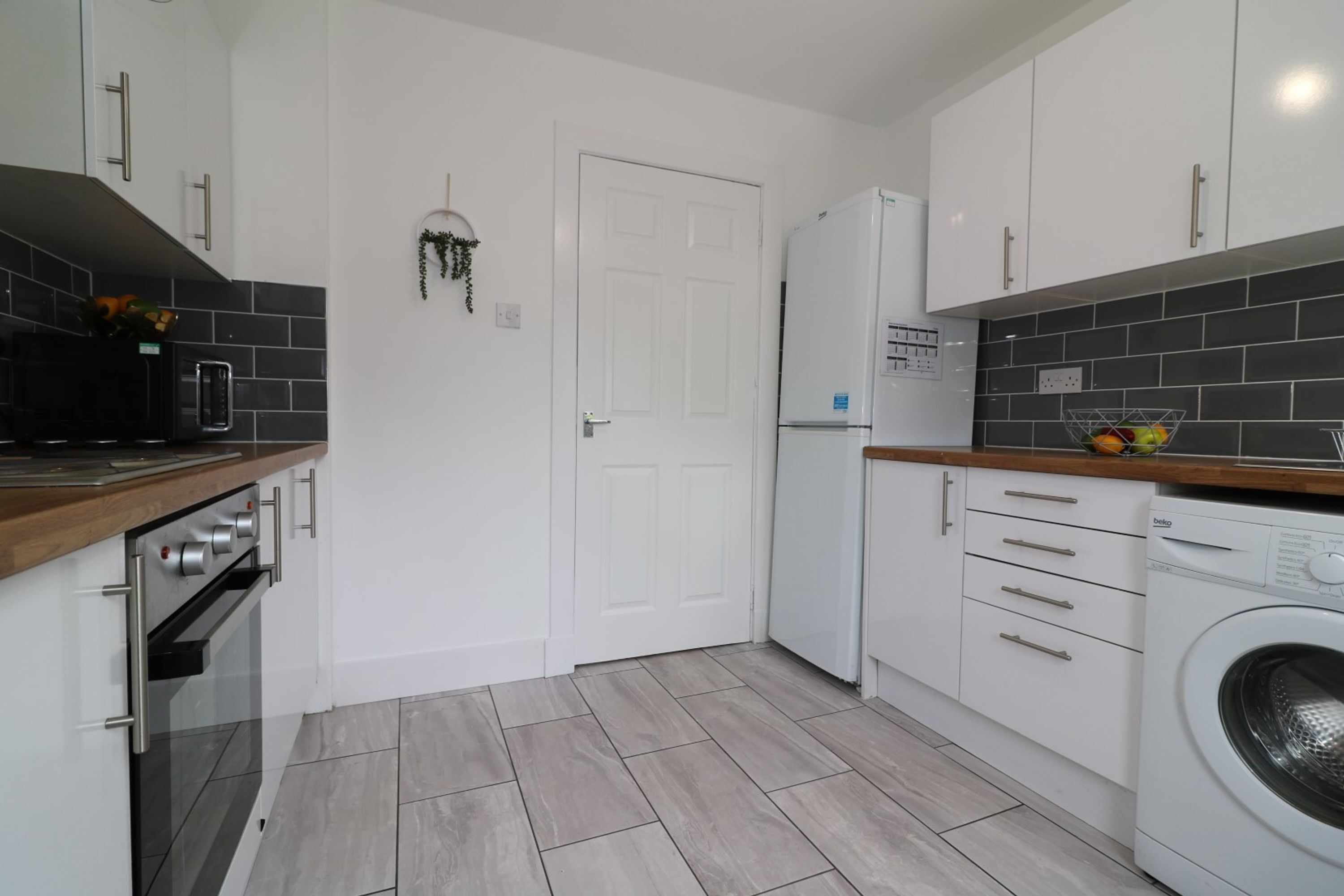 A compact kitchen with white cabinets, a built-in oven, a washing machine, and a small countertop with a fruit bowl.