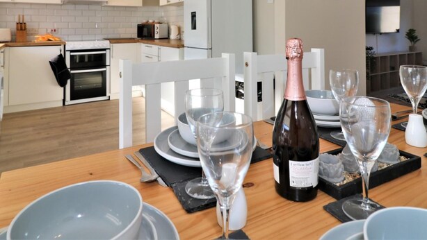 Una cocina moderna con armarios blancos, un comedor con mesa de madera dispuesta para una comida y una botella de champán.