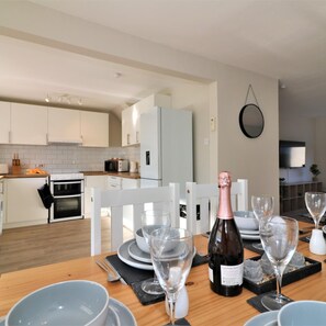 A modern kitchen with white cabinets, a dining area with a wooden table set for a meal, and a bottle of champagne. - Honeywell House - Airdrie (Airdrie)