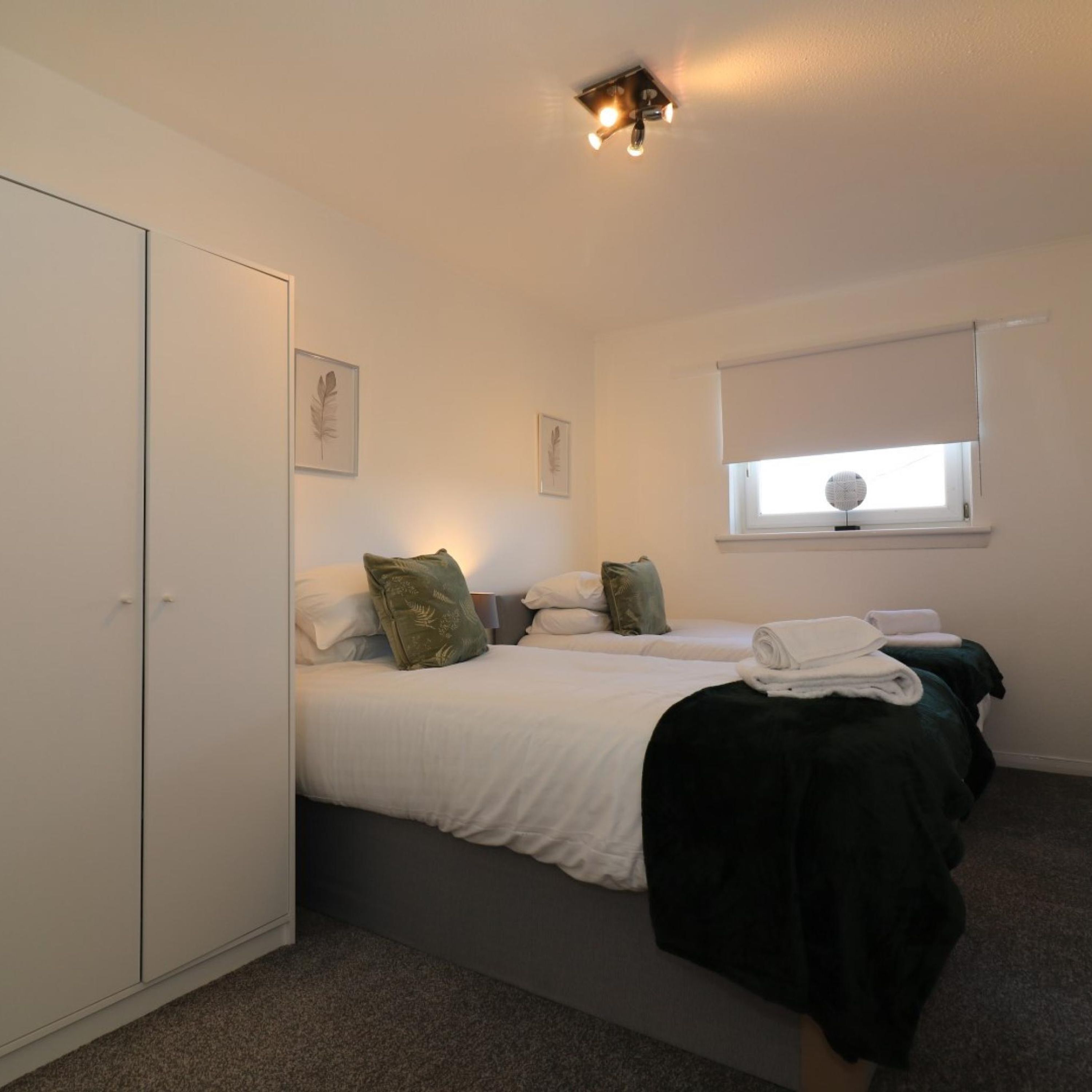 Une chambre avec deux lits, une armoire, une fenêtre avec des stores et un luminaire au plafond.