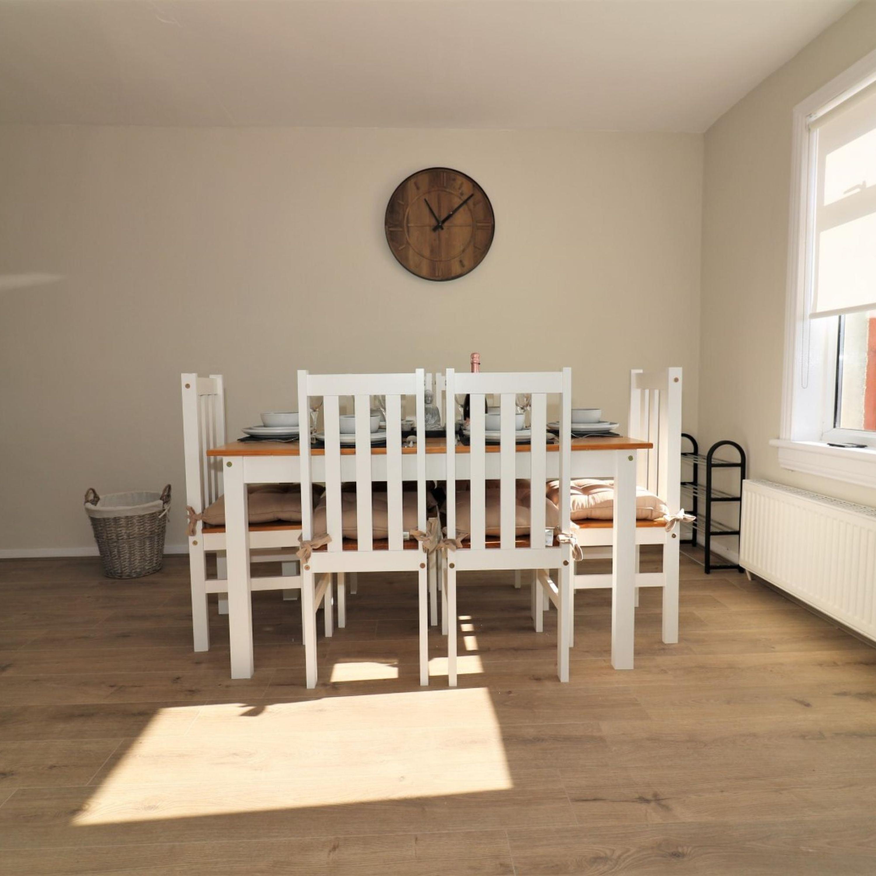 Een eetruimte met een houten tafel en witte stoelen, een klok aan de muur en een raam dat voor natuurlijk licht zorgt.