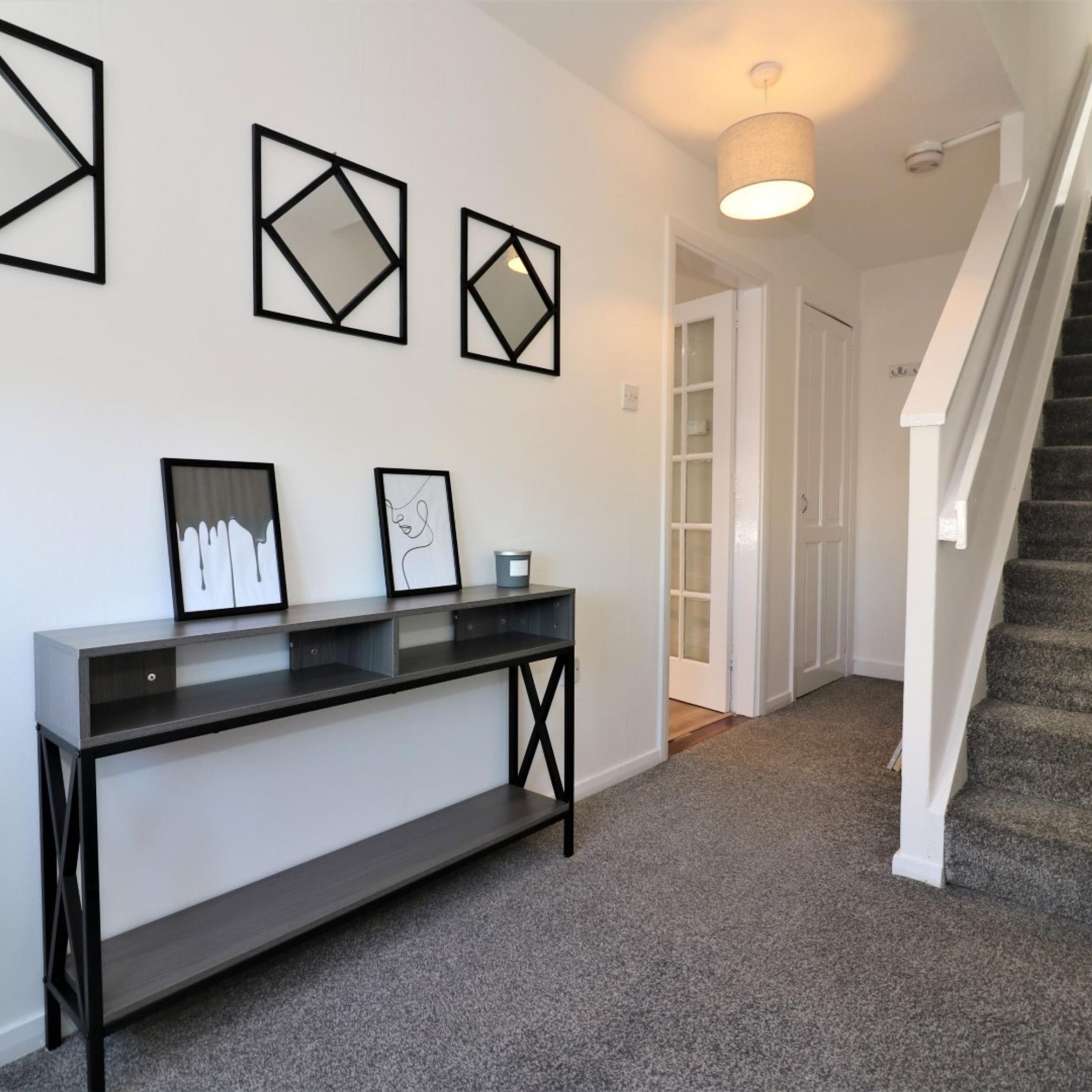 A hallway with a dark console table, three framed geometric wall art pieces, a white door, and a staircase with white railings.