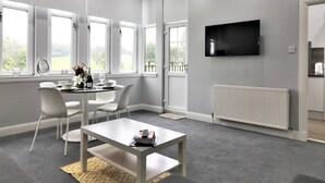 A modern living room with a grey carpet, white furniture, a flat-screen TV mounted on the wall, and large windows offering a view of greenery. - Greenlees Clubhouse 2 bed - Cambuslang (Glasgow)