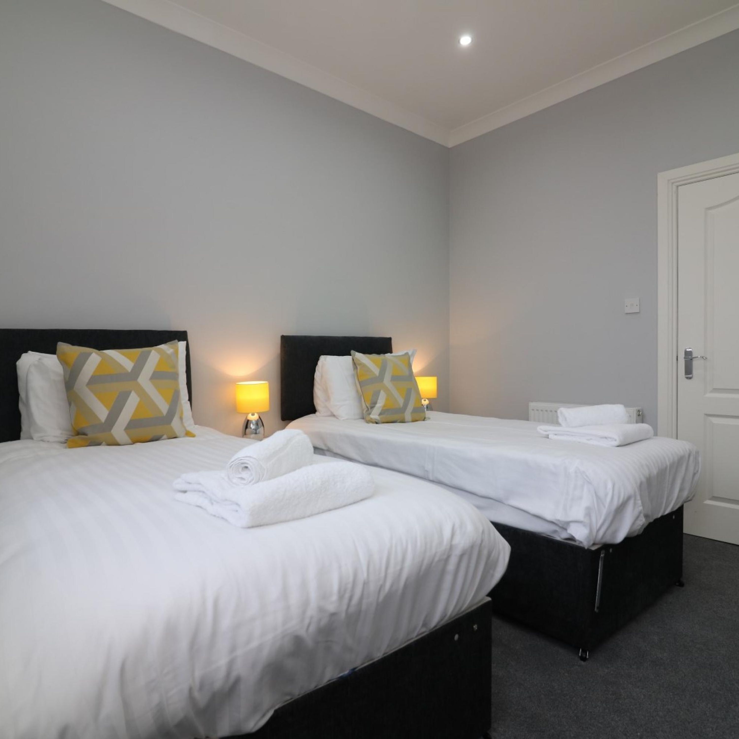 A hotel room with two single beds, each with a black headboard, white bedding, and a yellow and white patterned pillow. There are two bedside lamps with white shades, and a white door in the background.