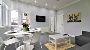A modern living room with a dining area, grey carpet, white furniture, a flat-screen TV, and a painting of donkeys. - Greenlees Clubhouse 2 bed - Cambuslang (Glasgow)