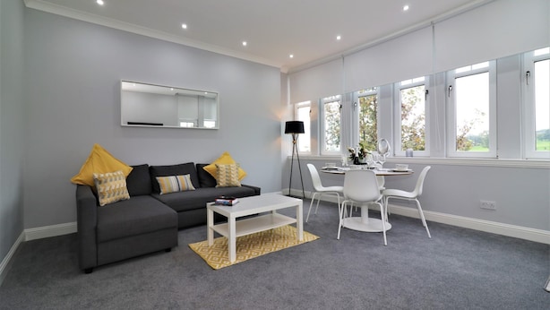 A modern living room with a grey sofa, white coffee table, and a dining area with white chairs. - Greenlees Clubhouse 2 bed - Cambuslang (Glasgow)