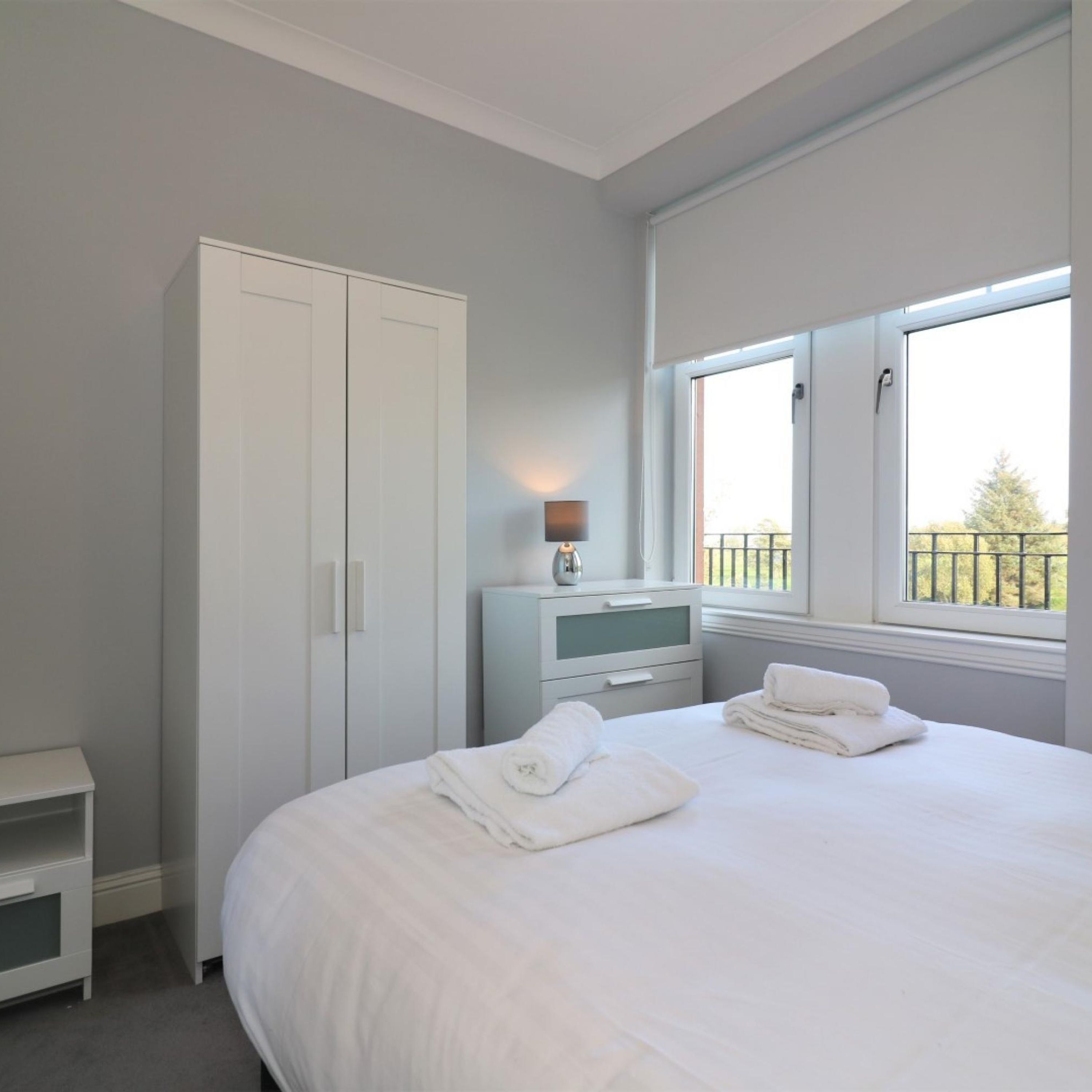 A bedroom with a large bed, a wardrobe, a bedside table, and a window with a view of greenery.