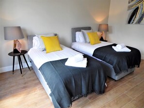 Een hotelkamer met twee bedden, elk met een zwart-wit dekbedovertrek, een geel kussen en een witte handdoek op het bed. Er staat een klein tafeltje met een lamp naast een van de bedden.