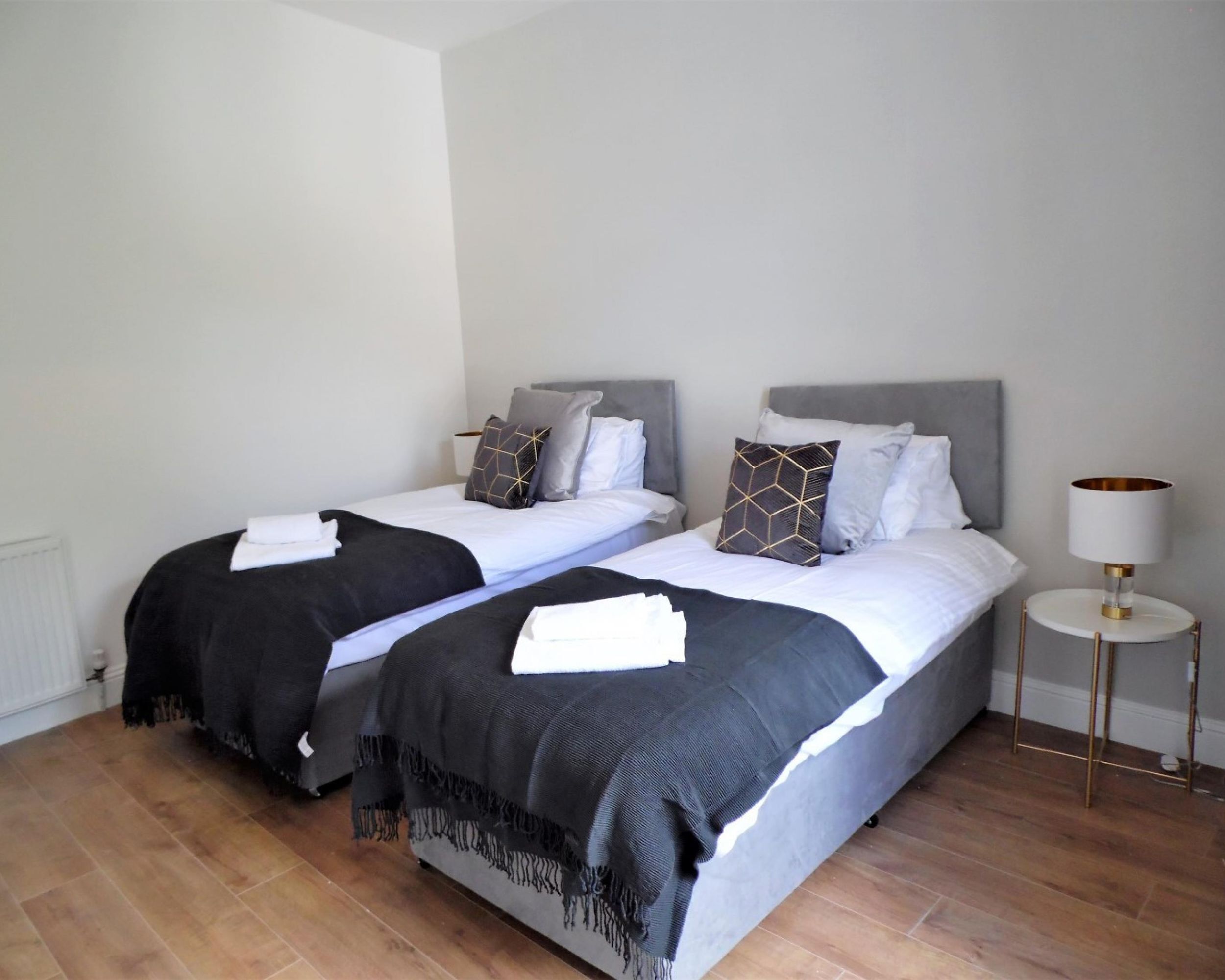 A double room with two beds, each with a grey headboard, white bedding, and decorative pillows. There is a bedside table with a lamp on the right side.