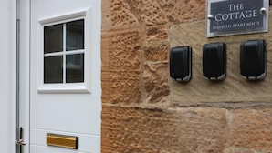 En vit dörr med ett fönster och ett mässingshandtag, en stenmur med en skylt som lyder ”The Cottage Serviced Apartments”, och tre svarta intercomsystem monterade på väggen.