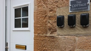 A white door with a window and a brass handle, a stone wall with a sign reading "The Cottage Serviced Apartments", and three black intercoms mounted on the wall. - Linden Cottage Room 1 - Airdrie (Airdrie)