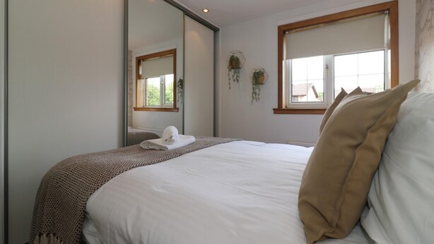Ein ordentlich bezogenes Bett mit weißer Decke und brauner Tagesdecke, ein großer Spiegel der den Raum reflektiert und ein Fenster mit Blick auf Gebäude.