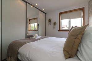 Een net opgemaakt bed met een witte deken en een bruine plaid, een grote spiegel die de kamer weerspiegelt, en een raam met uitzicht op gebouwen.