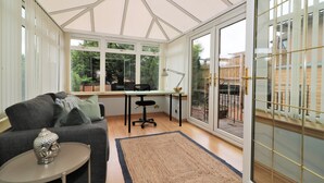 Ein Wintergarten mit einer Couch, einem Schreibtisch und einem Sessel, großen Fenstern und Blick ins Freie.