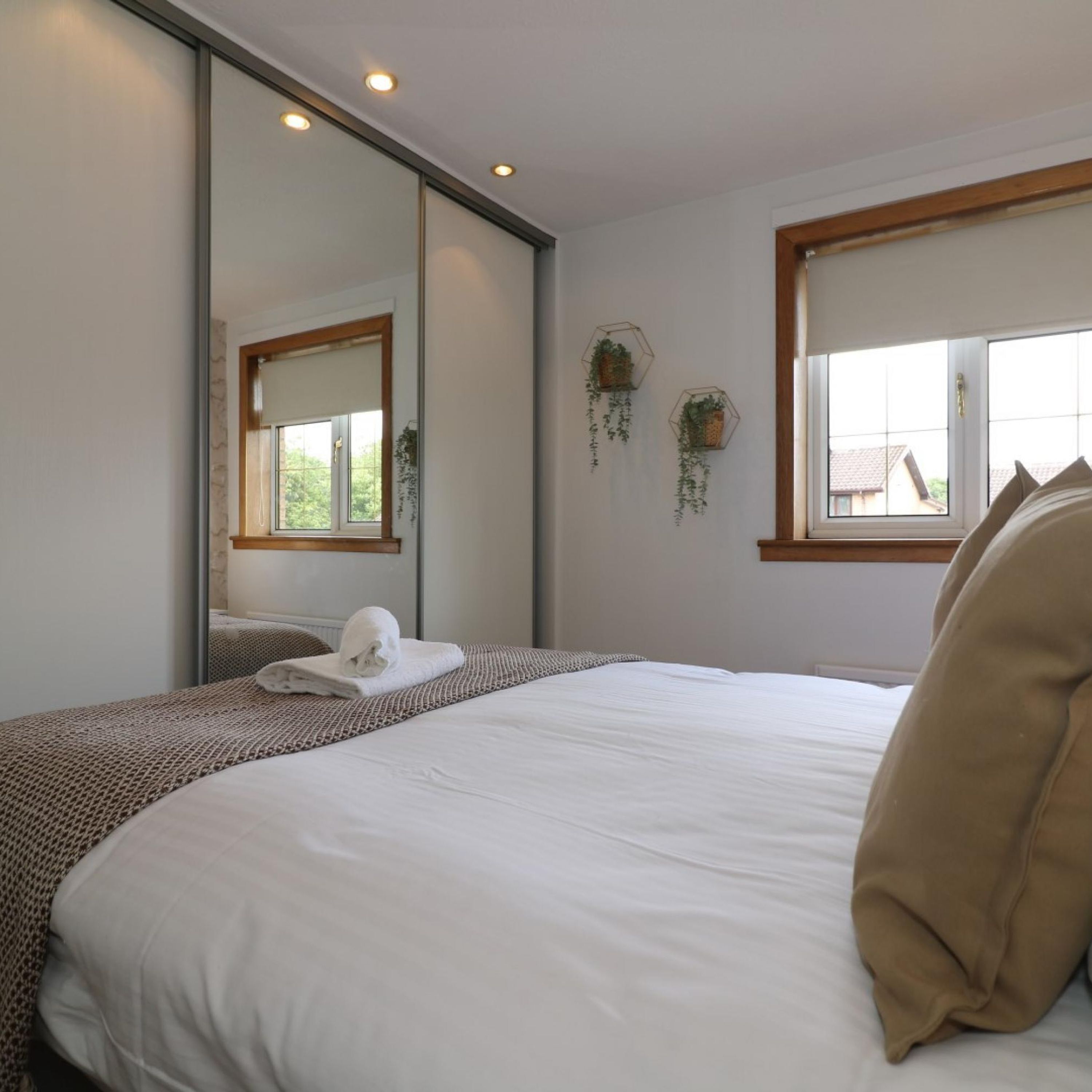 A hotel room with a large bed, a mirror, two windows, and hanging plants.