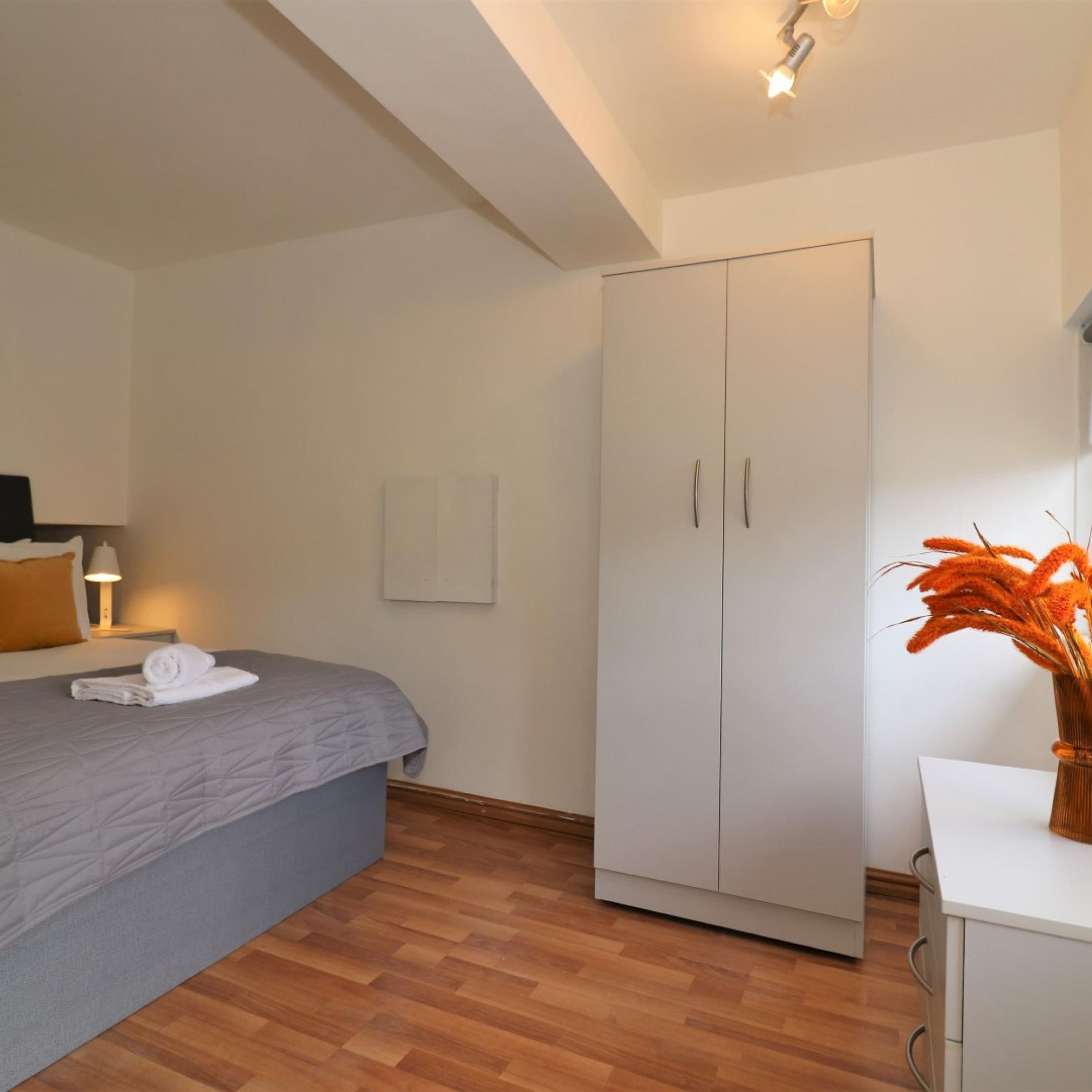 Une chambre à coucher avec un lit, une armoire, une table de chevet avec une lampe et un vase rempli de fleurs.