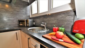 A modern kitchen with a stainless steel sink, a microwave, and a red appliance. There are fresh vegetables on a wooden cutting board. - Linden Cottage Room 4 - Airdrie (Airdrie)