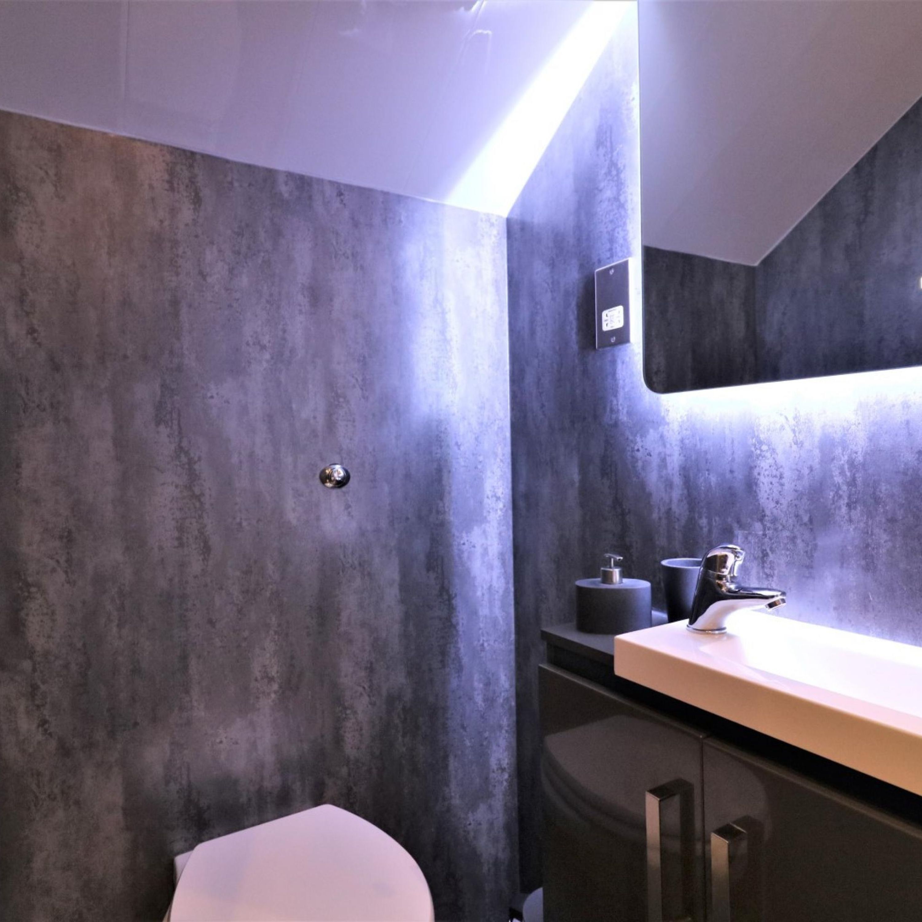 A modern bathroom with a grey textured wall, a white toilet, a sink with a chrome faucet, and a small mirror above the sink.