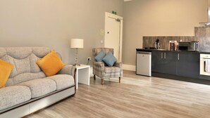 A modern living room with a sofa, a chair, a small table, and a kitchen area with a microwave and a coffee maker. - Linden House - Flat 3 - Airdrie (Airdrie)