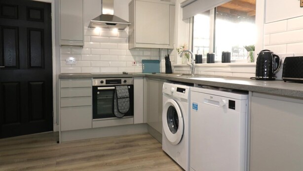 A modern kitchen with white cabinetry, a built-in oven, a washing machine, and a window providing natural light. - Sunnyside House - Holytown (Motherwell)
