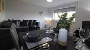 A modern living room with a dining table set for two, a sofa, a small side table, and a potted plant. - Sunnyside House - Holytown (Motherwell)