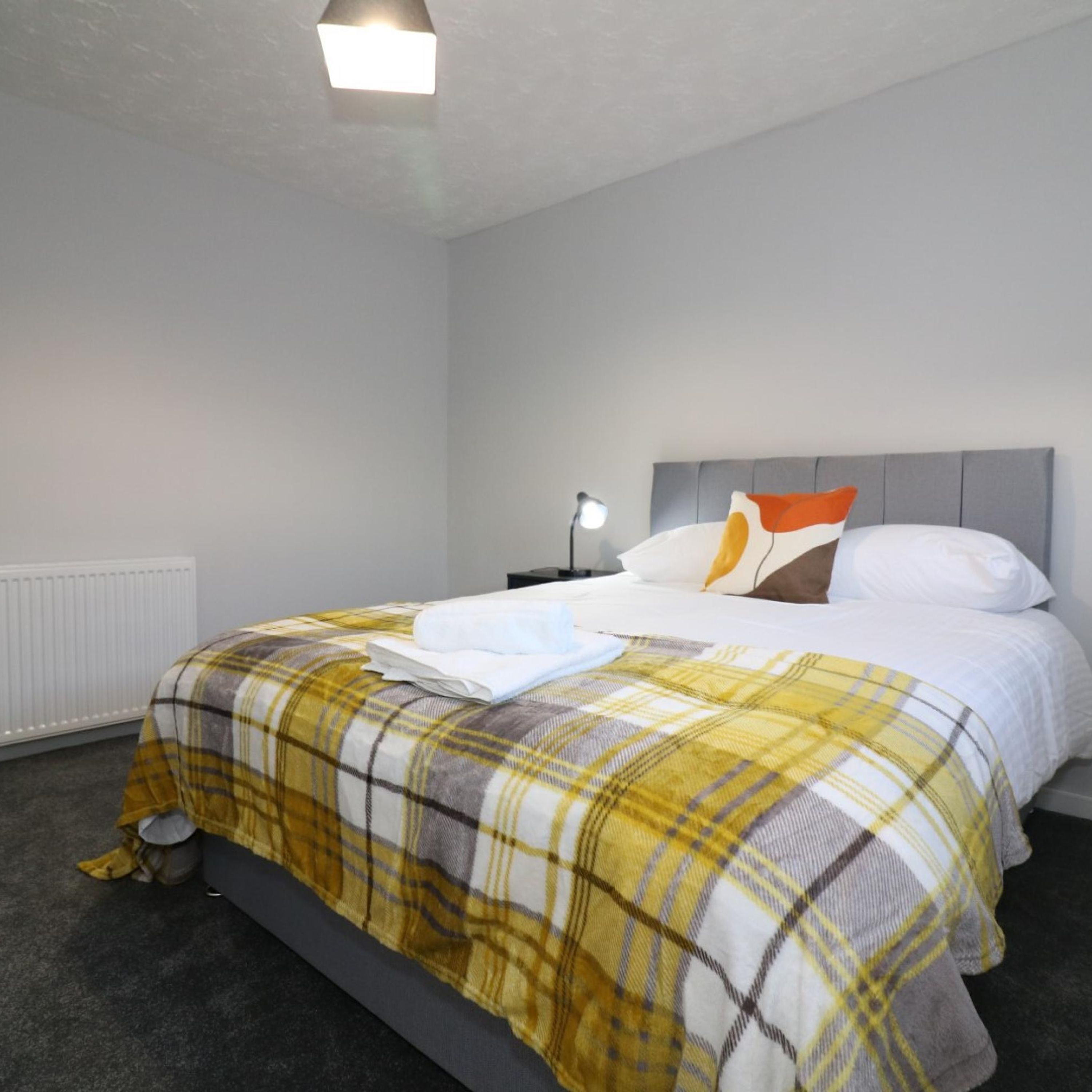 A neatly made bed with a plaid bedspread, a bedside lamp, and a radiator on the wall.