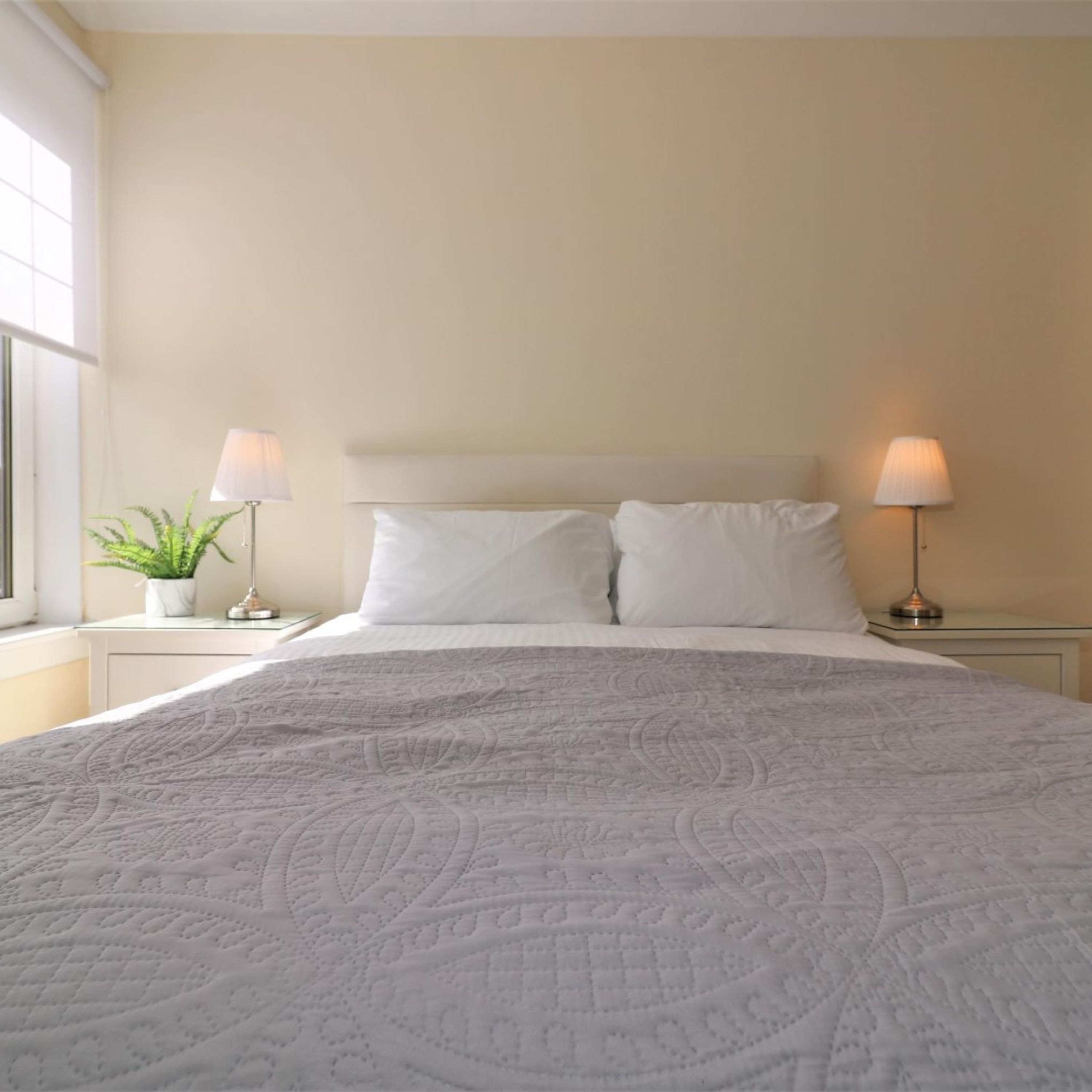 Una cama bien hecha con una colcha estampada, dos lámparas de noche, una planta en maceta y una ventana a la izquierda.