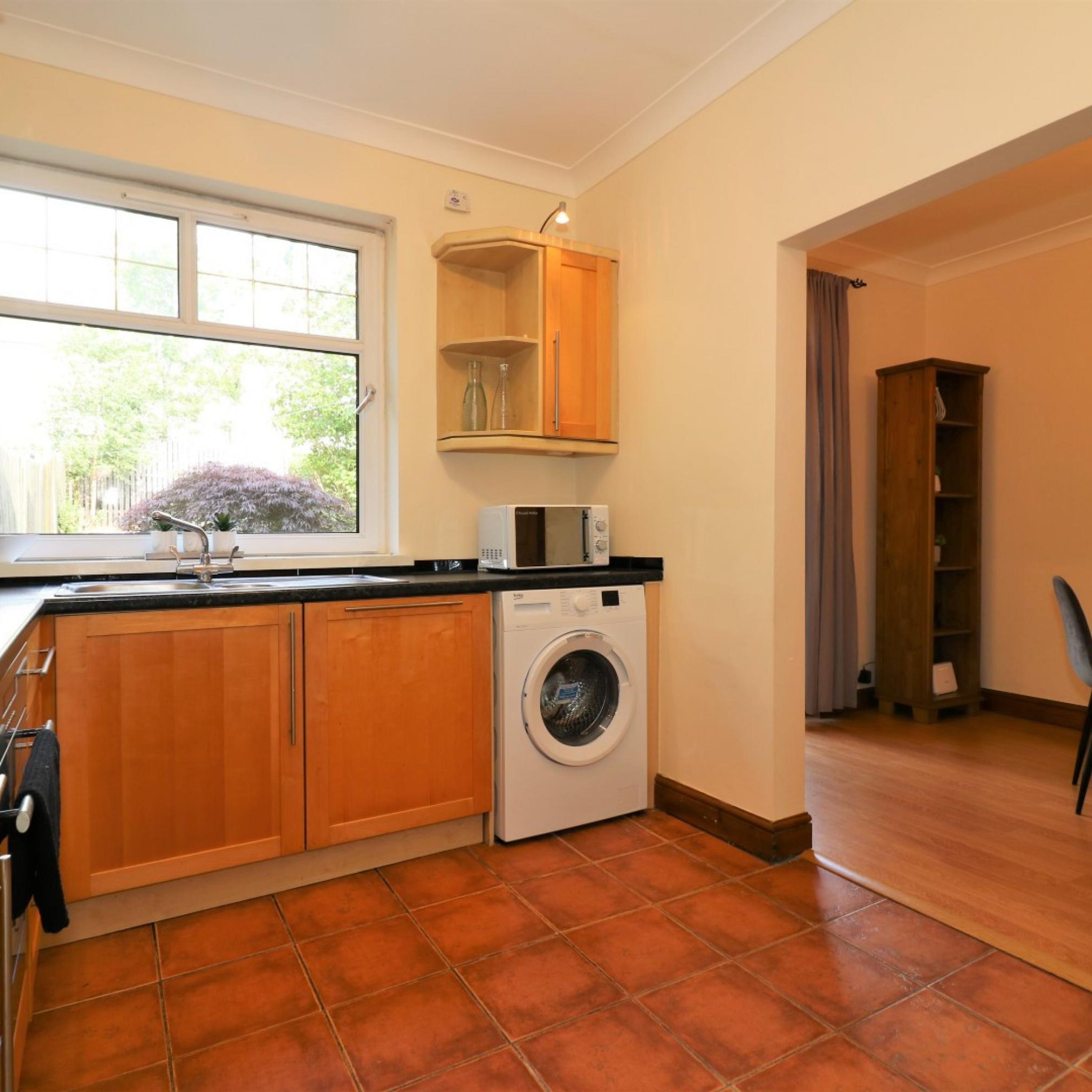 A compact kitchen with wooden cabinets, a washing machine, a microwave, and a window with a view of greenery.