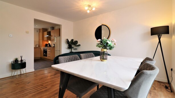A modern dining area with a white table, dark cushioned chairs, a vase with flowers, a round mirror, and a floor lamp. - Belvidere House - Parkhead (Glasgow)