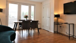 A modern living room with a dining area, wooden flooring, a flat-screen TV mounted on the wall, and a ceiling light fixture. - Belvidere House - Parkhead (Glasgow)