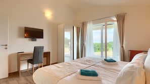 Quarto de hotel com cama, escrivaninha, cadeira, televisão e varanda com cortinas.