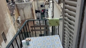 Un petit balcon avec une table recouverte d’une nappe à motifs, un coussin vert et deux verres.