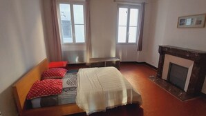 Une chambre à coucher avec un lit, deux fenêtres, un banc, une cheminée et un tableau au mur.