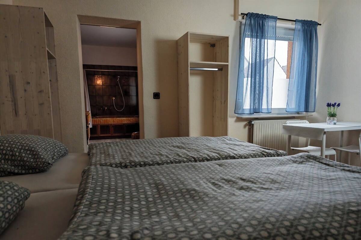 Quarto com duas camas, um guarda-roupa de madeira, uma mesinha e uma janela com cortinas azuis.