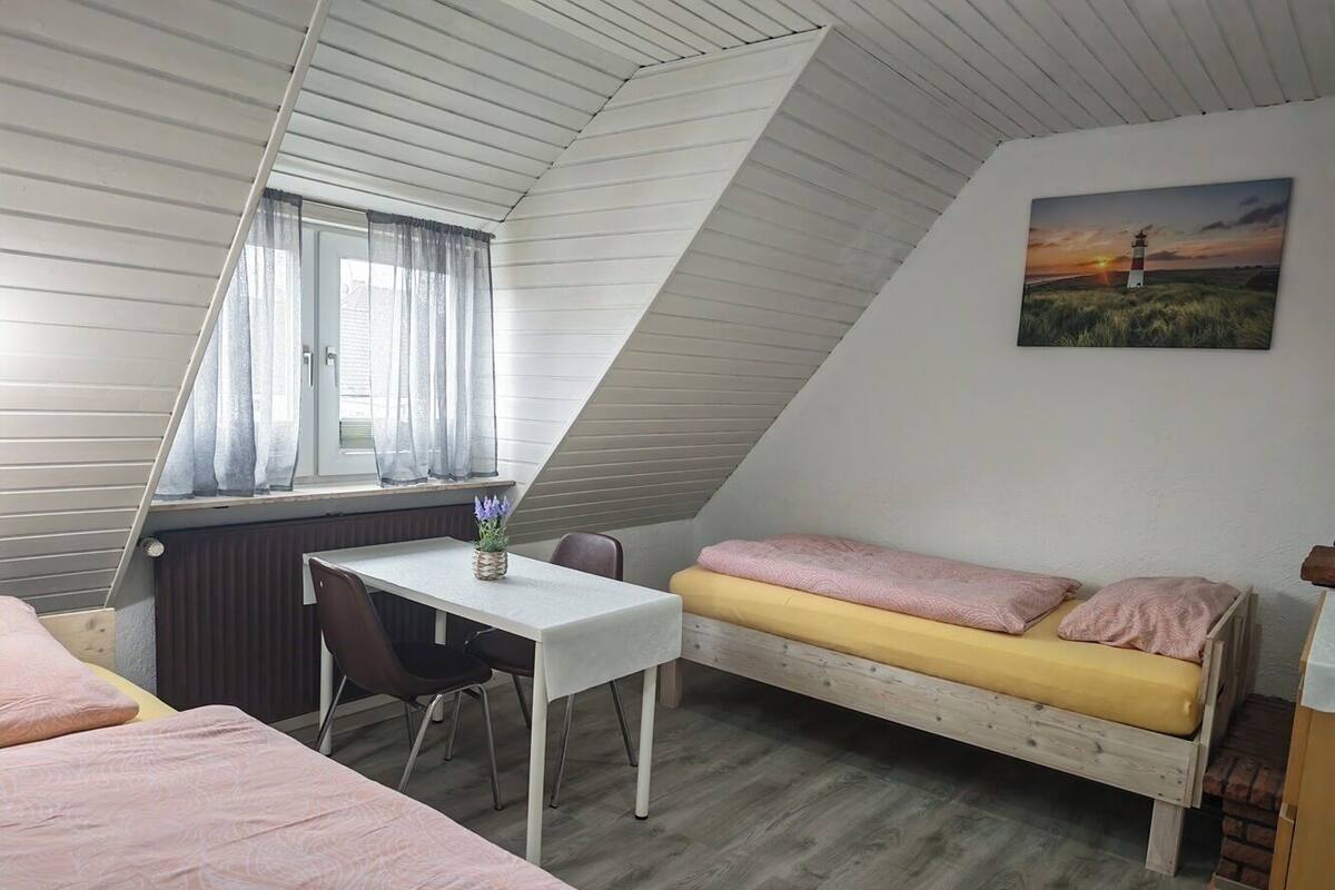 Quarto em dormitório com teto inclinado, uma cama, uma escrivaninha e uma janela.