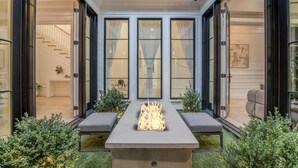 A modern outdoor patio with a fire feature, seating, and a view of the interior through large windows.