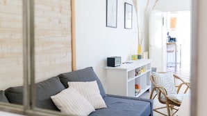 A modern living room with a grey sofa, white cushions, a white shelving unit with books and decorative items, and a wicker chair. - Holiday home quartier Arago Les Sables - 8 people (Les Sables-d´Olonne)