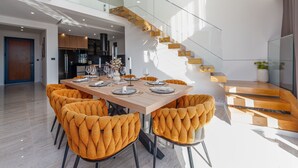 A modern dining area with a wooden table set for six, surrounded by tufted orange chairs. A glass staircase leads to an upper level, and a kitchen is visible in the background.