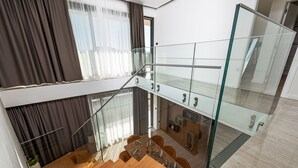 A modern interior with a glass staircase connecting two floors, a dining area with wooden furniture, and a view of the outdoors through large windows.