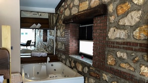 A modern bathroom with a large bathtub, stone walls, and a view of a balcony with outdoor seating. - MASKLAVİ (Kestel)