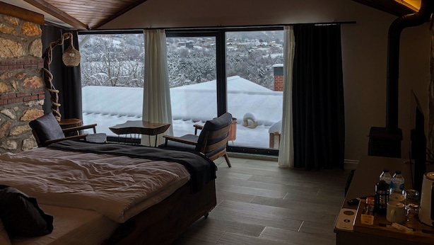 A bedroom with a large bed, a wooden ceiling, a stone wall, a fireplace, a window with curtains, and a view of a snowy landscape. - MASKLAVİ (Kestel)