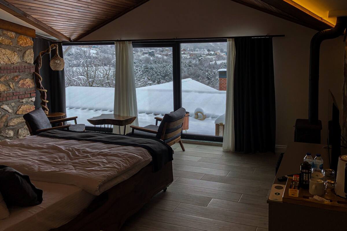 A bedroom with a large bed, a wooden ceiling, a stone wall, a fireplace, a window with curtains, and a view of a snowy landscape.