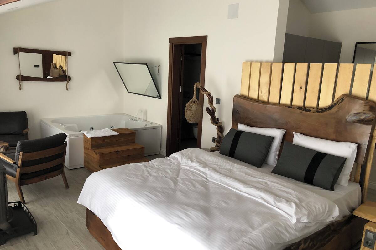 A bedroom with a large bed, a wooden headboard, a wooden chair, a small desk, a hanging basket, and a bathroom with a bathtub and sink.