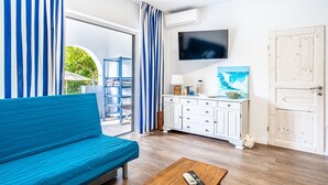 Een woonkamer met een blauwe bank, een houten salontafel, een aan de muur bevestigde televisie en een wit kastje met een schilderij.