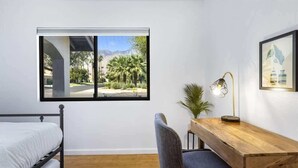 A bedroom with a bed, a desk, a chair, a lamp, a window with a view of palm trees, and a framed picture on the wall.